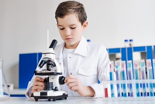 preteen boy in white coat near microscope and blurred test tubes in lab