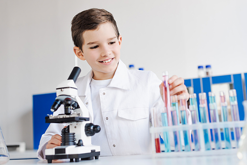 smiling preteen boy in white coat holding test tube near microscope in chemical laboratory