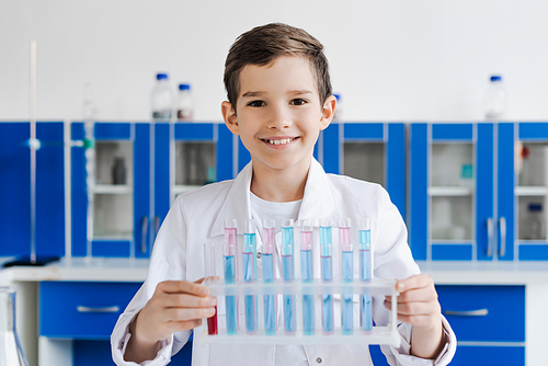 cheerful boy in white coat looking at camera near test tubes in laboratory