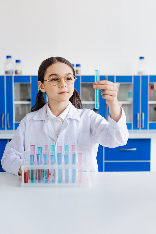 preteen girl in eyeglasses and white coat looking at test tube in chemical laboratory