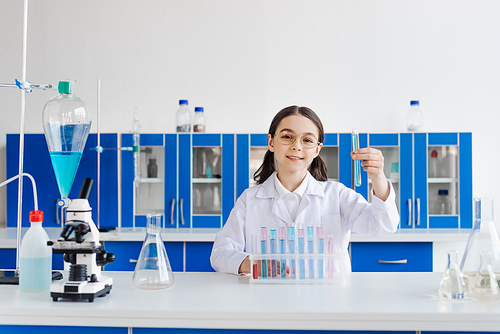 cheerful girl in eyeglasses and white coat holding test tube while sitting near microscope and flasks