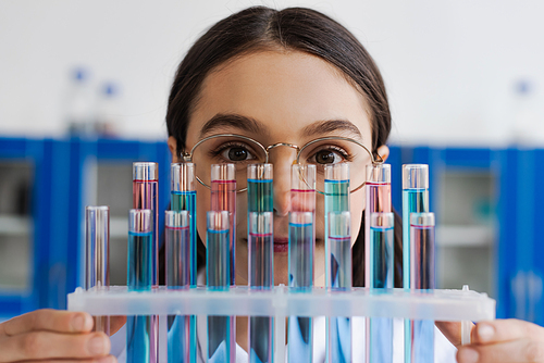 brunette girl in eyeglasses looking at camera near test tubes with liquid