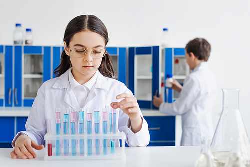 girl in white coat and eyeglasses near test tubes with liquid and boy on blurred background