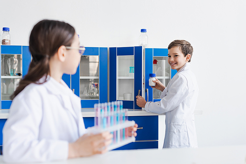 smiling boy in white coat holding jar with powder near girl with test tubes on blurred foreground