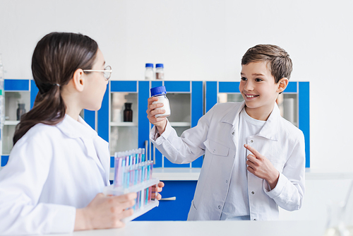 happy boy pointing at jar with white substance near blurred girl with test tubes
