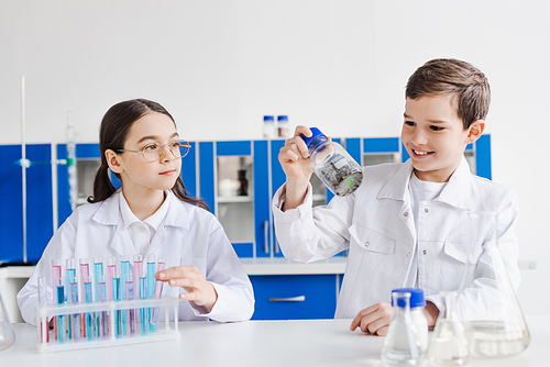 cheerful boy holding jar with chemical substance near girl in eyeglasses and test tubes in lab