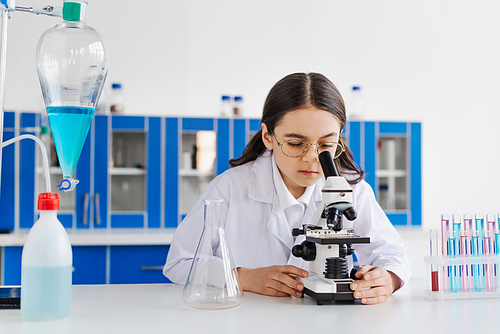preteen girl in eyeglasses looking in microscope near test tubes and flasks