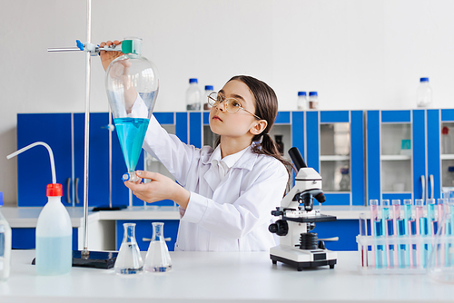 preteen girl in eyeglasses and white coat holding flask near microscope and test tubes