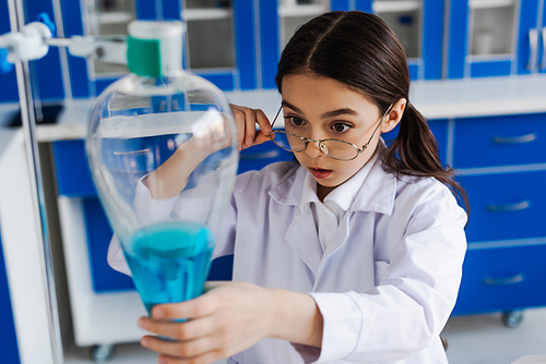 preteen girl holding eyeglasses while looking at blurred flask with blue liquid during chemical experiment
