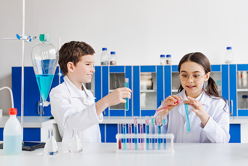 happy preteen kids in white coats looking at camera near microscope and test tubes with flasks