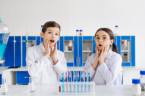 shocked preteen kids in white coats touching faces and looking at camera in chemical laboratory