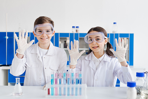 smiling kids in white coats and goggles showing hands in latex gloves near test tubes in lab