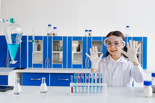 smiling girl in goggles showing hands in latex gloves near flasks and test tubes in lab