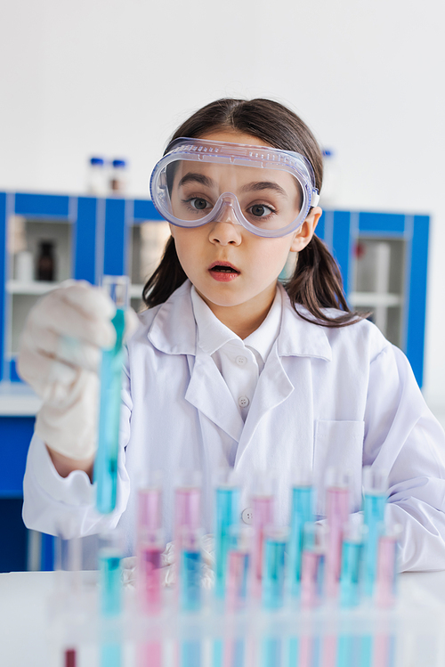 amazed girl in white coat and goggles looking at blurred test tube in lab