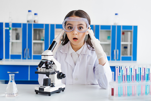 amazed girl in goggles and latex gloves looking at camera near microscope and test tubes