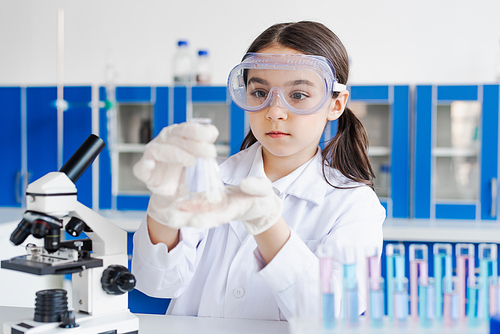 girl in goggles holding flask near microscope in laboratory