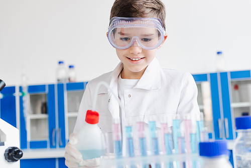 smiling boy in goggles holding bottle near blurred test tubes during chemical experiment in laboratory