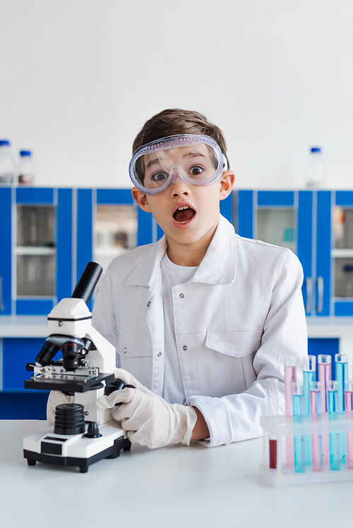amazed boy in goggles looking at camera near microscope and test tubes in laboratory