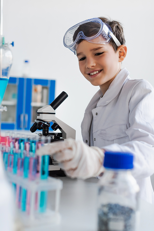 cheerful boy with goggles smiling near blurred test tubes and microscope