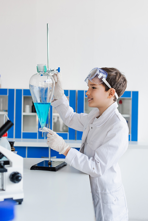 side view of smiling kid in latex gloves looking at flask with blue liquid near microscope