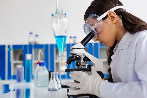 side view of preteen girl in goggles looking into microscope in chemical laboratory