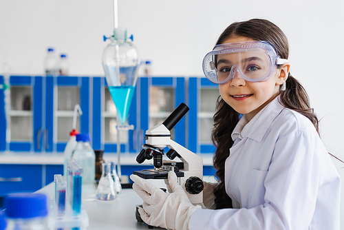 preteen girl in goggles and white coat smiling at camera near microscope in lab