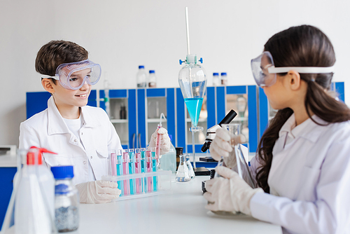 smiling boy in goggles looking at blurred girl near test tubes and flasks in lab