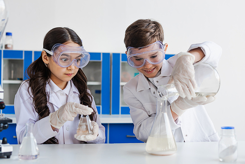 tensed girl in goggles looking at smiling boy pouring liquid into flask while doing chemical experiment