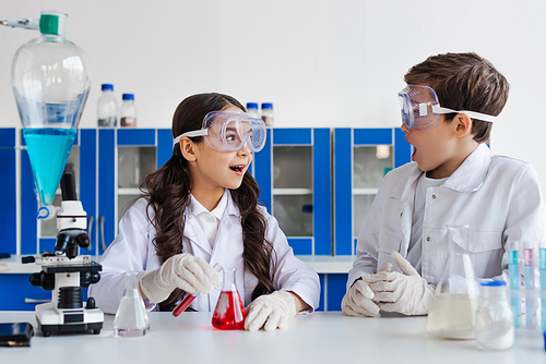 amazed girl in goggles looking at friend near flask with red liquid in chemical lab