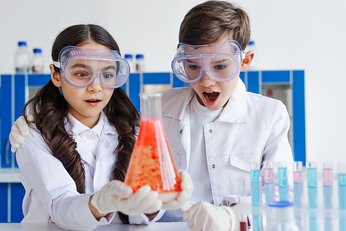 amazed friends in goggles looking at flask with red substance in laboratory