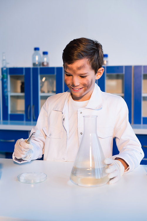 smiling boy with dirty face holding tweezers near petri dish and flask in lab
