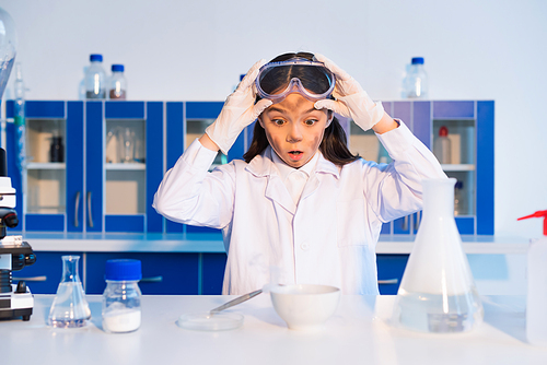 shocked girl with dirty face holding goggles and looking at bowl during chemical experiment