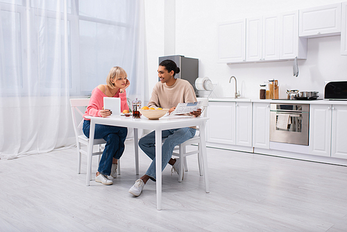 happy african american man holding newspaper near blonde woman with digital tablet during breakfast