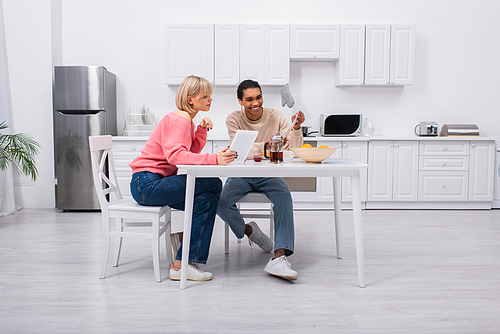 cheerful african american man holding newspaper near blonde woman with digital tablet during breakfast