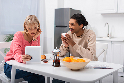 happy african american man looking at blonde woman with digital tablet during breakfast