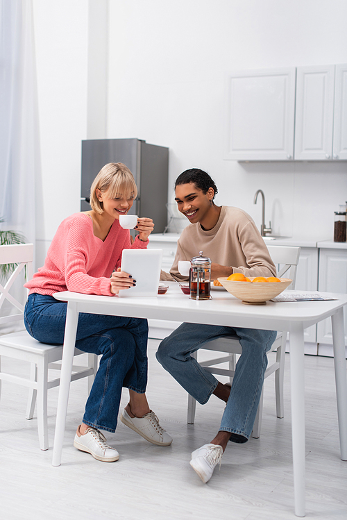 happy african american man and cheerful woman looking at digital tablet during breakfast