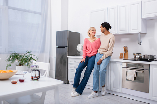 full length of happy multiethnic couple looking at each other in modern kitchen