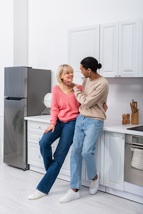 full length of smiling multiethnic couple looking at each other in modern kitchen