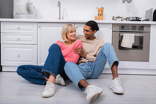 cheerful multiethnic couple sitting on floor in modern kitchen