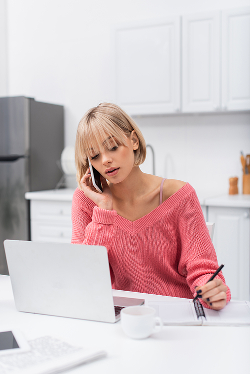 young freelancer talking on smartphone while writing near laptop