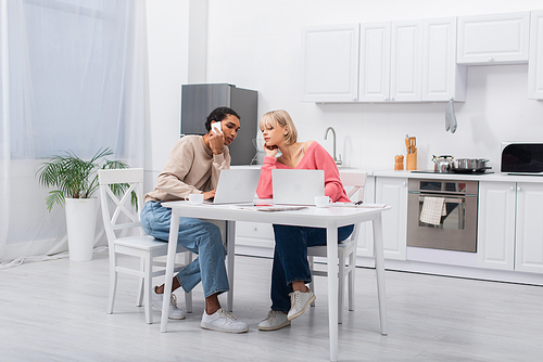 young african american freelancer talking on smartphone near blonde girlfriend and laptops