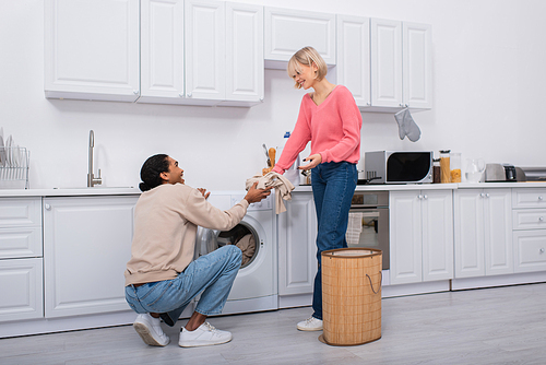 happy blonde woman passing dirty clothes to cheerful african american man