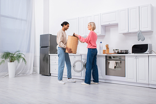 african american man holding basket near washing machine and happy girlfriend