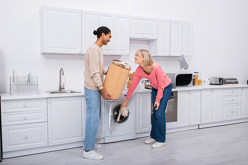 happy blonde woman putting clothes in washing machine near african american man