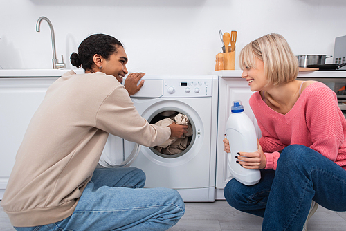 happy blonde woman holding bottle with detergent near african american man putting clothes in washing machine