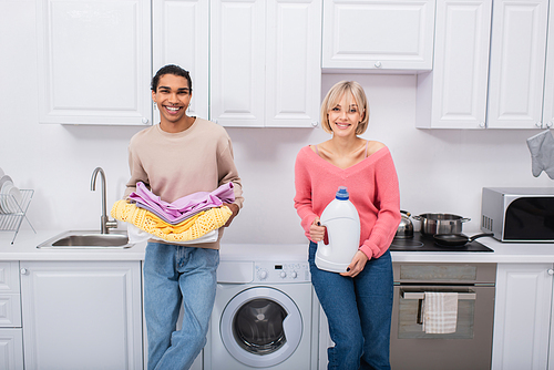 happy interracial couple holding bottle with detergent and colorful clothes