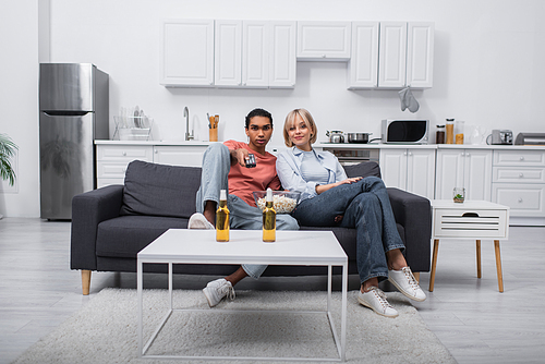 african american man holding remote controller and clicking channels near blonde girlfriend in living room