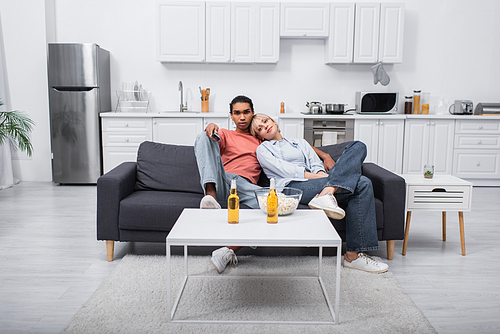 young blonde woman leaning on african american man holding remote controller in living room