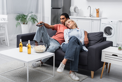 blonde woman leaning on african american boyfriend holding remote controller in living room