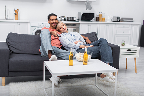 young blonde woman leaning on happy african american man holding remote controller in living room